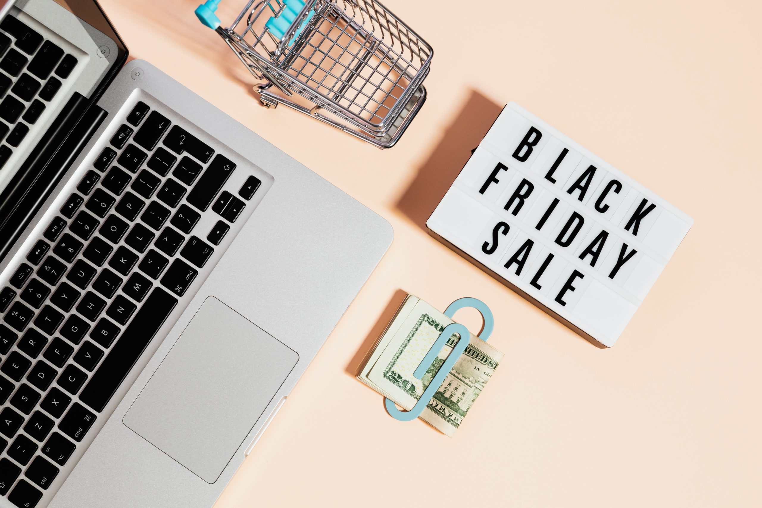 Un MacBook, un caddie miniature, un billet de banque et un message affichant "Black Friday Sale" sur un fond orange clair.