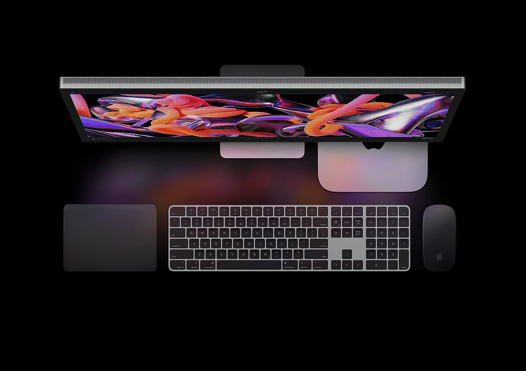 Set-up Apple composé d'un écran, d'un Mac Mini M2, d'un trackpad, d'un clavier et d'une souris. Le fond de l'image est uni et sombre.
